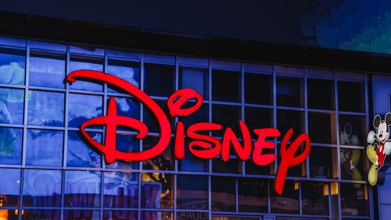 The image features a vibrant storefront of a Disney retail location, prominently displaying the iconic Disney logo in red against a glass façade. Part of the storefront includes a colorful depiction of Mickey Mouse.