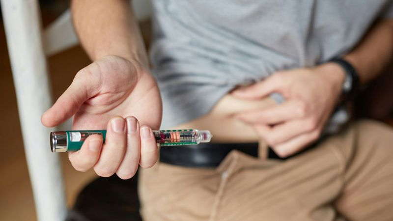 The image shows a person preparing to administer an injection using a pen-style injector device. The individual is seated and has lifted their shirt to expose their abdomen, indicating the area for the injection.