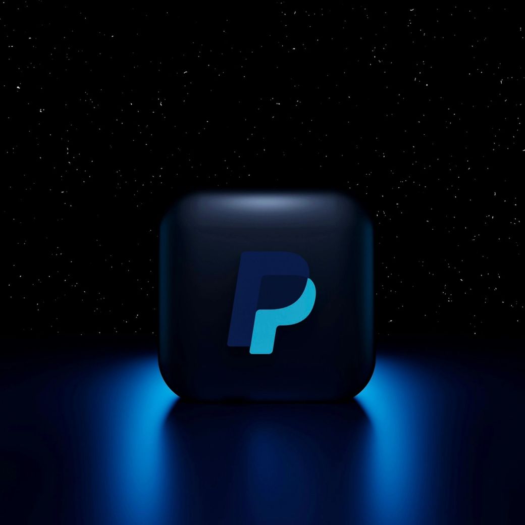 The image features a dark, glowing representation of the PayPal logo set against a starry background. The logo is highlighted in shades of blue, suggesting a modern and digital aesthetic. — Credit: Photo by Mariia Shalabaieva / Unsplash