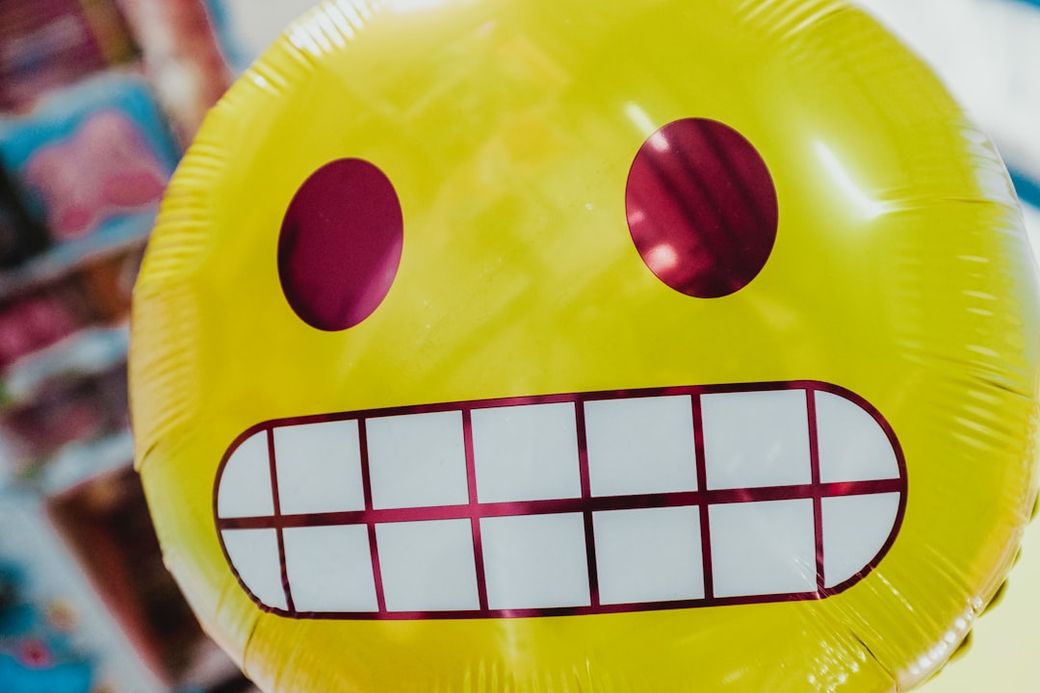 The image features a vibrant yellow balloon with a large, cheerful smiley face design. The balloon stands out against a blurred background, suggesting a festive atmosphere, possibly in a party or celebration setting. — Credit: Photo by Bernard Hermant on Unsplash