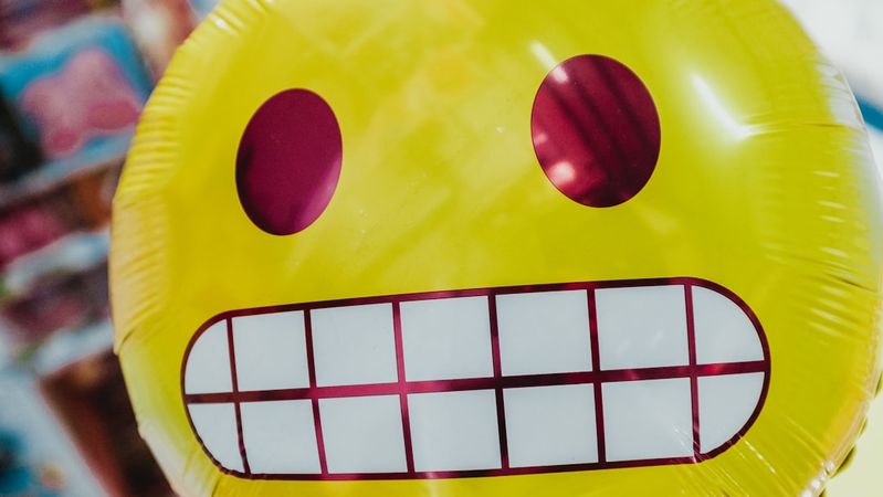 The image features a vibrant yellow balloon with a large, cheerful smiley face design. The balloon stands out against a blurred background, suggesting a festive atmosphere, possibly in a party or celebration setting.
