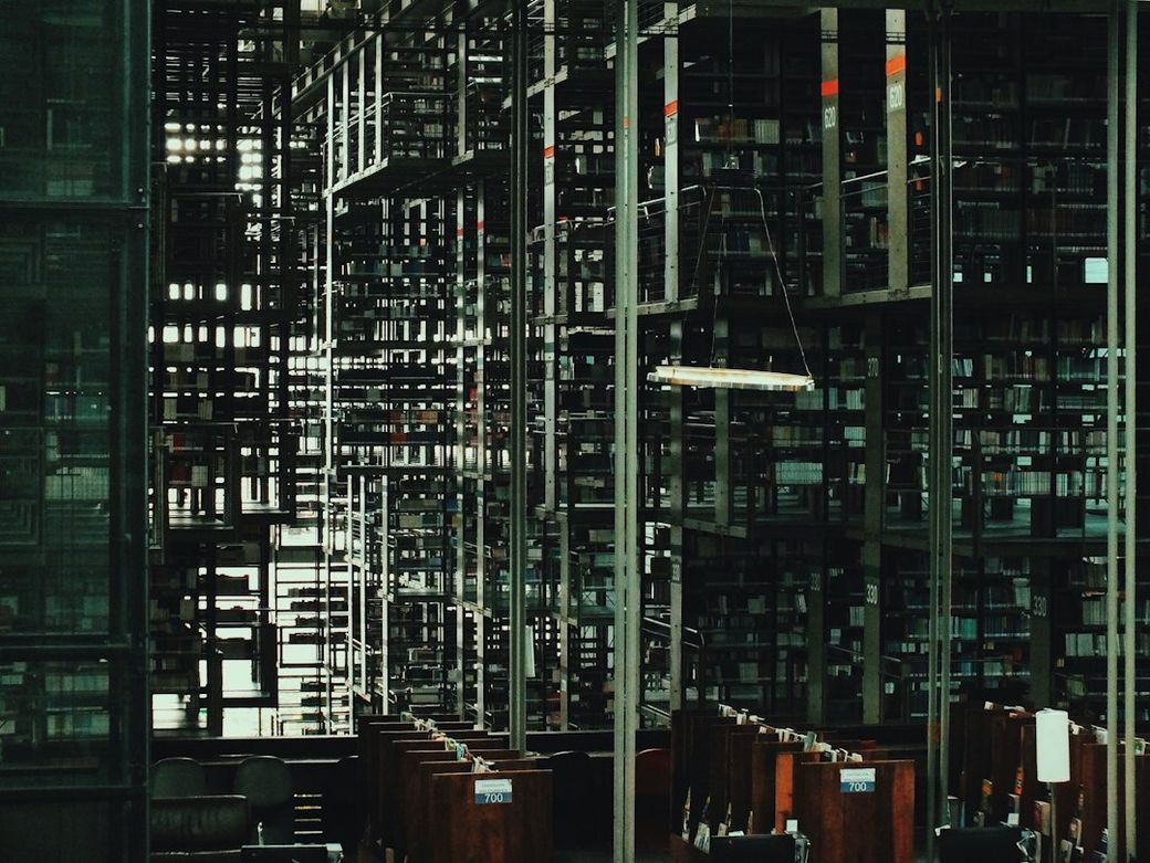 The image showcases a modern library interior characterized by expansive metal shelving filled with books. The arrangement of shelves and seating creates a maze-like effect, highlighting the interplay of light and shadow. — Credit: Photo by Guillermo Ruiz on Unsplash