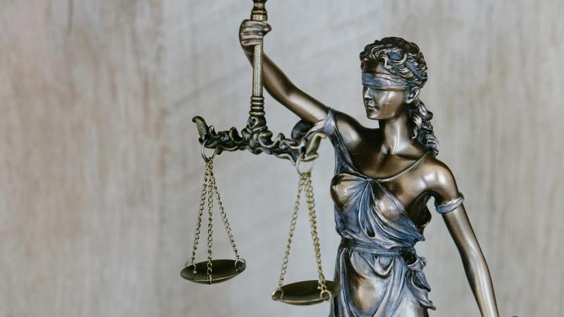 The image features a bronze statue of Lady Justice, symbolizing fairness and law. She is depicted blindfolded, holding a scale in one hand and a sword in the other.