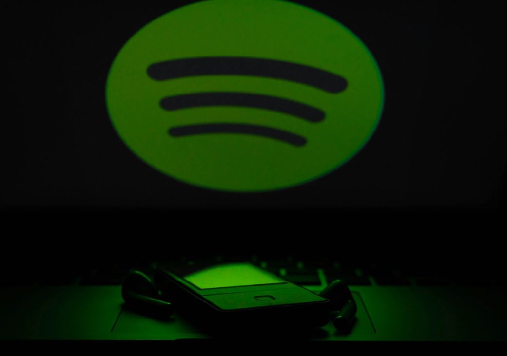The image features a green Spotify logo prominently displayed against a dark background, with a blurred foreground of a device, possibly a headset or smartphone. The logo illuminates the surroundings, creating a contrast with the dark environment. — Credit: Photo by Reet Talreja / Unsplash