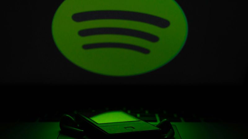 The image features a green Spotify logo prominently displayed against a dark background, with a blurred foreground of a device, possibly a headset or smartphone. The logo illuminates the surroundings, creating a contrast with the dark environment.