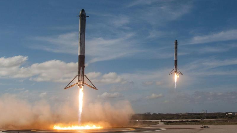 The image captures two rockets launching simultaneously from a launch pad, leaving trails of fire and smoke. The backdrop features a partly cloudy sky, emphasizing the power and technology behind the launch.