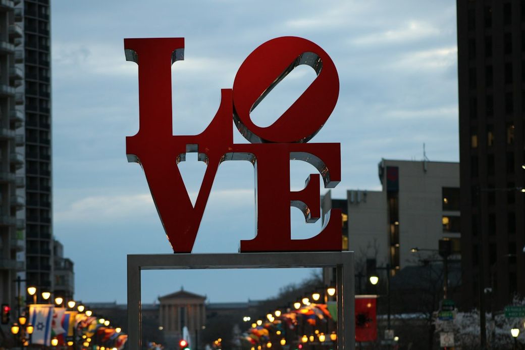 The image features a large, iconic 'LOVE' sculpture prominently displayed in an urban setting, with trees and buildings in the background. The architecture reflects a modern cityscape under an overcast sky. — Credit: Photo by Salonagility.com Daniel on Unsplash