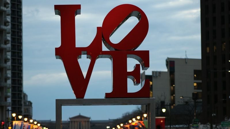 The image features a large, iconic 'LOVE' sculpture prominently displayed in an urban setting, with trees and buildings in the background. The architecture reflects a modern cityscape under an overcast sky.