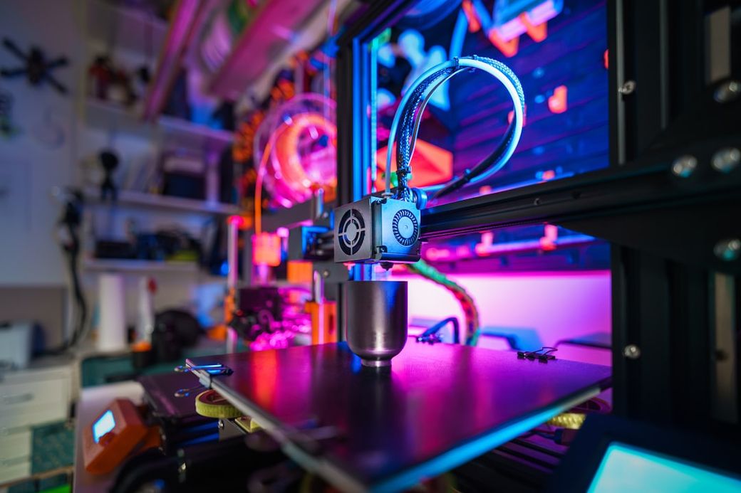 The image shows a close-up of a 3D printer in operation, featuring colorful lighting that highlights its intricate machinery. The print bed is visible, showcasing the printing process with a metallic object being created. — Credit: Photo by Jakub Żerdzicki on Unsplash