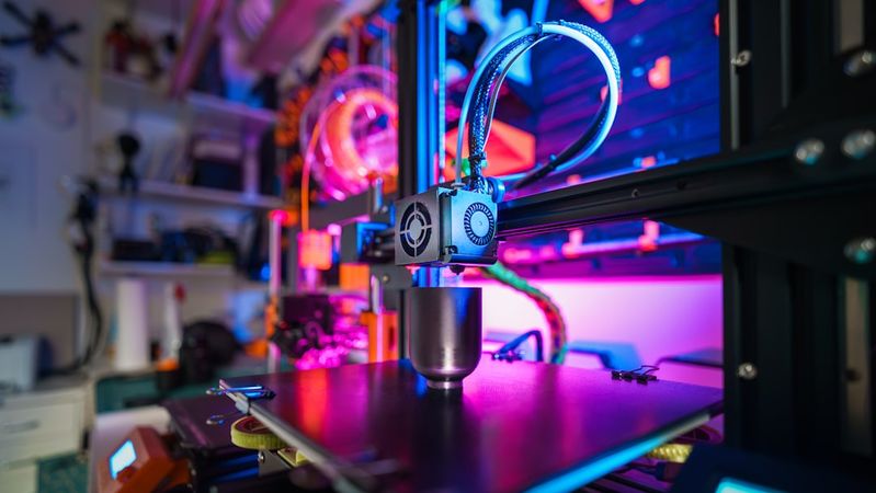 The image shows a close-up of a 3D printer in operation, featuring colorful lighting that highlights its intricate machinery. The print bed is visible, showcasing the printing process with a metallic object being created.