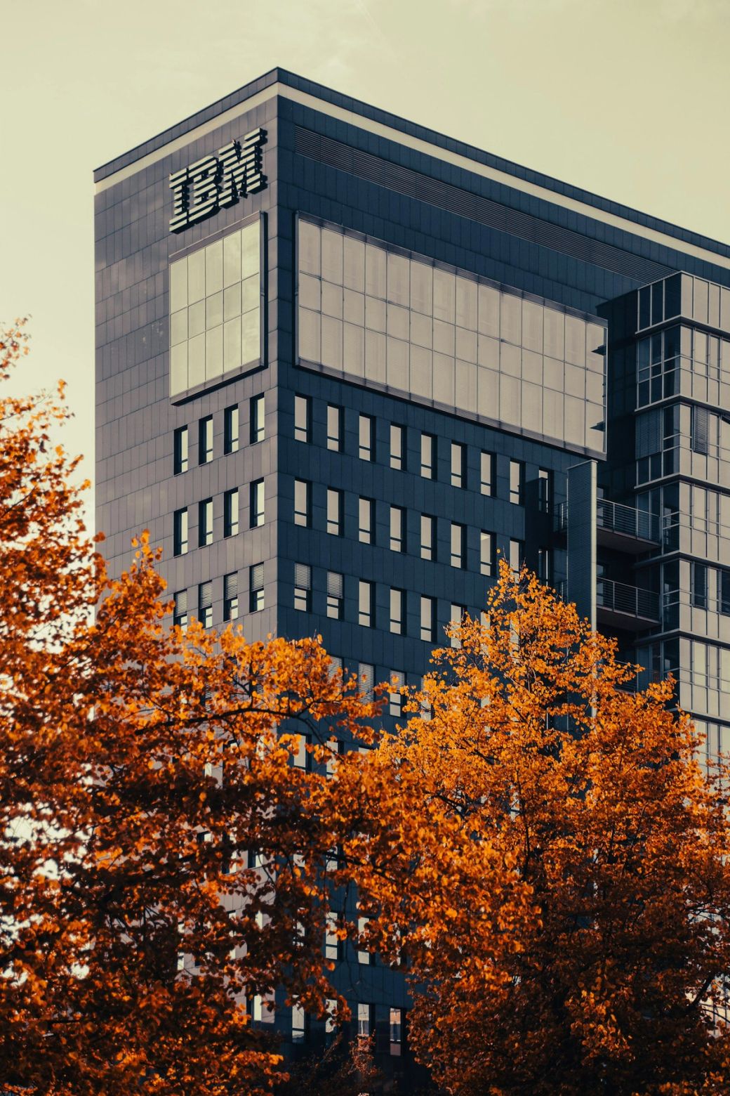 The image features the top of a modern office building with a prominent IBM logo displayed on its facade. The architectural design reflects a sleek and contemporary style, indicating a corporate environment. — Credit: Photo by Denny Müller / Unsplash