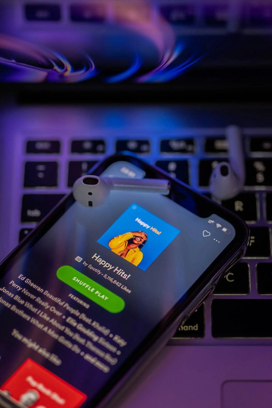 A smartphone displaying the 'Happy Hits!' playlist on the Spotify app, with a pair of wireless earbuds resting nearby. The image is taken against a dark background, highlighting the phone's screen and the app's vibrant interface. — Credit: Photo by omid armin / Unsplash