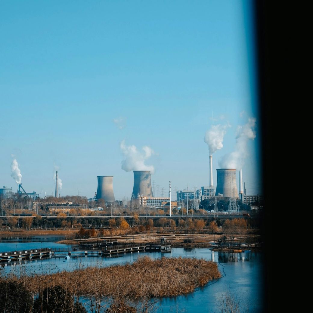 The image depicts a skyline featuring several industrial cooling towers emitting white steam against a clear blue sky. In the foreground, there are signs of an industrial area with infrastructure visible. — Credit: Photo by Meatball Overexposure / Unsplash