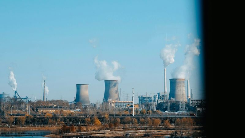 The image depicts a skyline featuring several industrial cooling towers emitting white steam against a clear blue sky. In the foreground, there are signs of an industrial area with infrastructure visible.