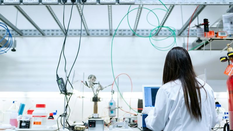 The image shows a laboratory setting where a researcher, wearing a white lab coat, is focused on a laptop. The workspace features various equipment and supplies, highlighting a scientific environment.