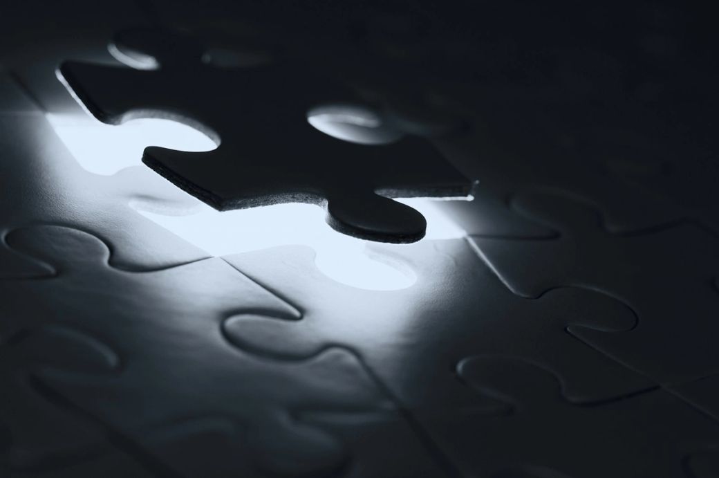 The image features a close-up of a puzzle with a piece missing, illuminated by light coming through the gap. The surrounding puzzle pieces are in shadow, emphasizing the absence of the missing piece. — Credit: Photo by Edge2Edge Media / Unsplash