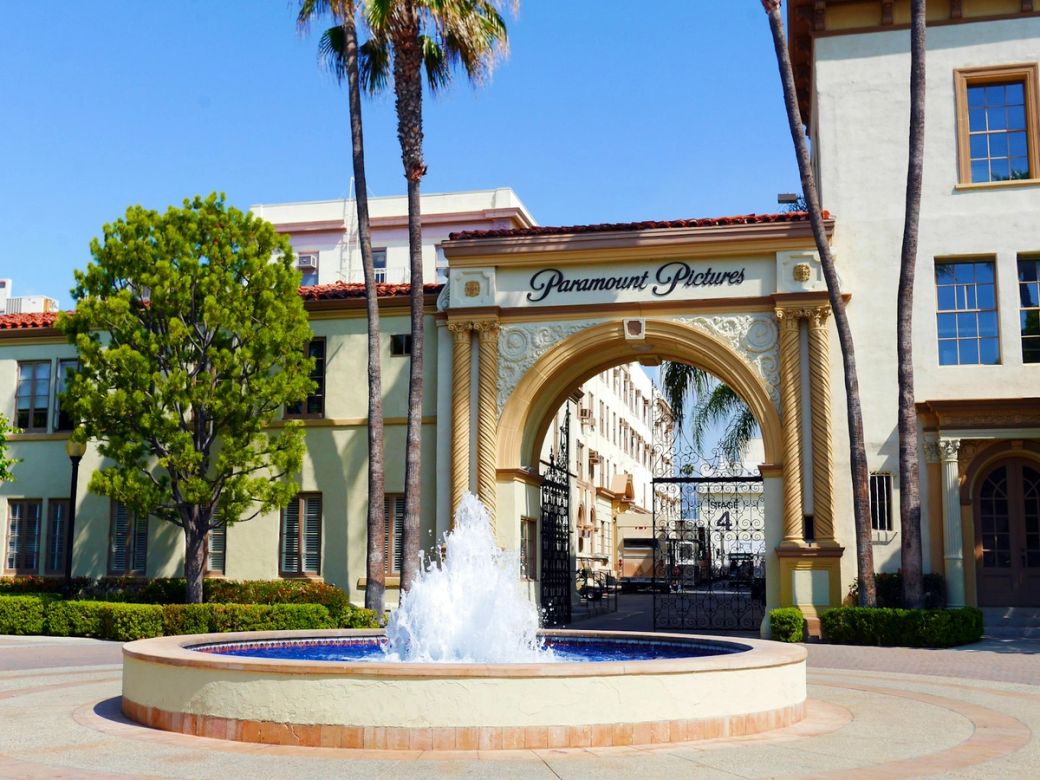 The image depicts the entrance archway of Paramount Pictures surrounded by a landscaped area featuring palm trees and a fountain. The iconic branding of Paramount Pictures is prominently displayed above the entrance. — Credit: Photo by Hannah Wernecke / Unsplash
