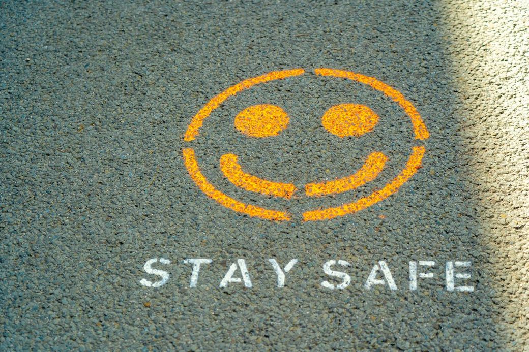 The image shows a smiley face graphic along with the text 'STAY SAFE' painted on a textured asphalt surface. The bright orange and white colors stand out against the gray background, conveying a positive and reassuring message. — Credit: Photo by Nick Fewings on Unsplash