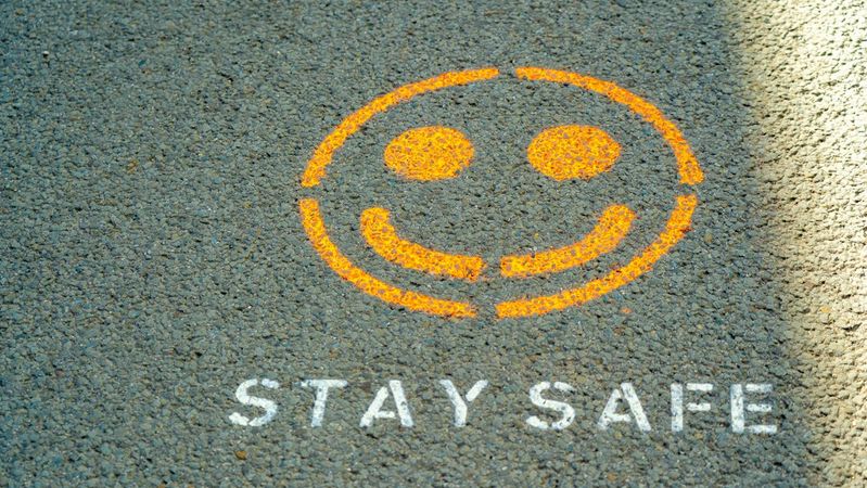 The image shows a smiley face graphic along with the text 'STAY SAFE' painted on a textured asphalt surface. The bright orange and white colors stand out against the gray background, conveying a positive and reassuring message.