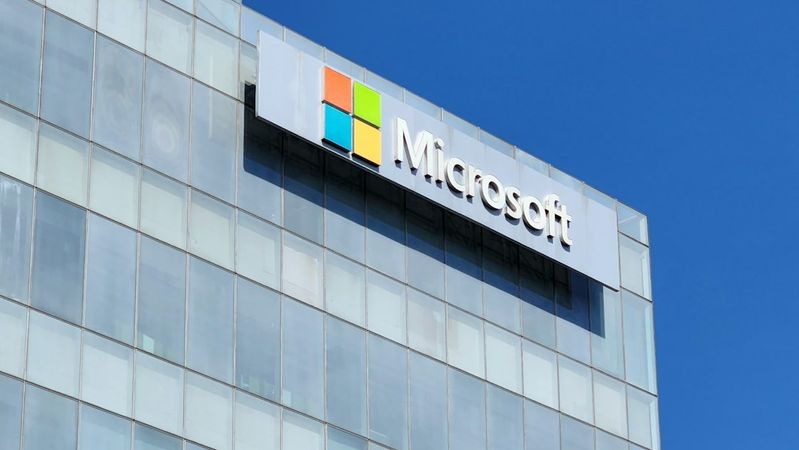 The image features the exterior of a modern office building with a prominent Microsoft logo displayed on the facade. The bright blue sky serves as a contrasting backdrop to the glass structure.