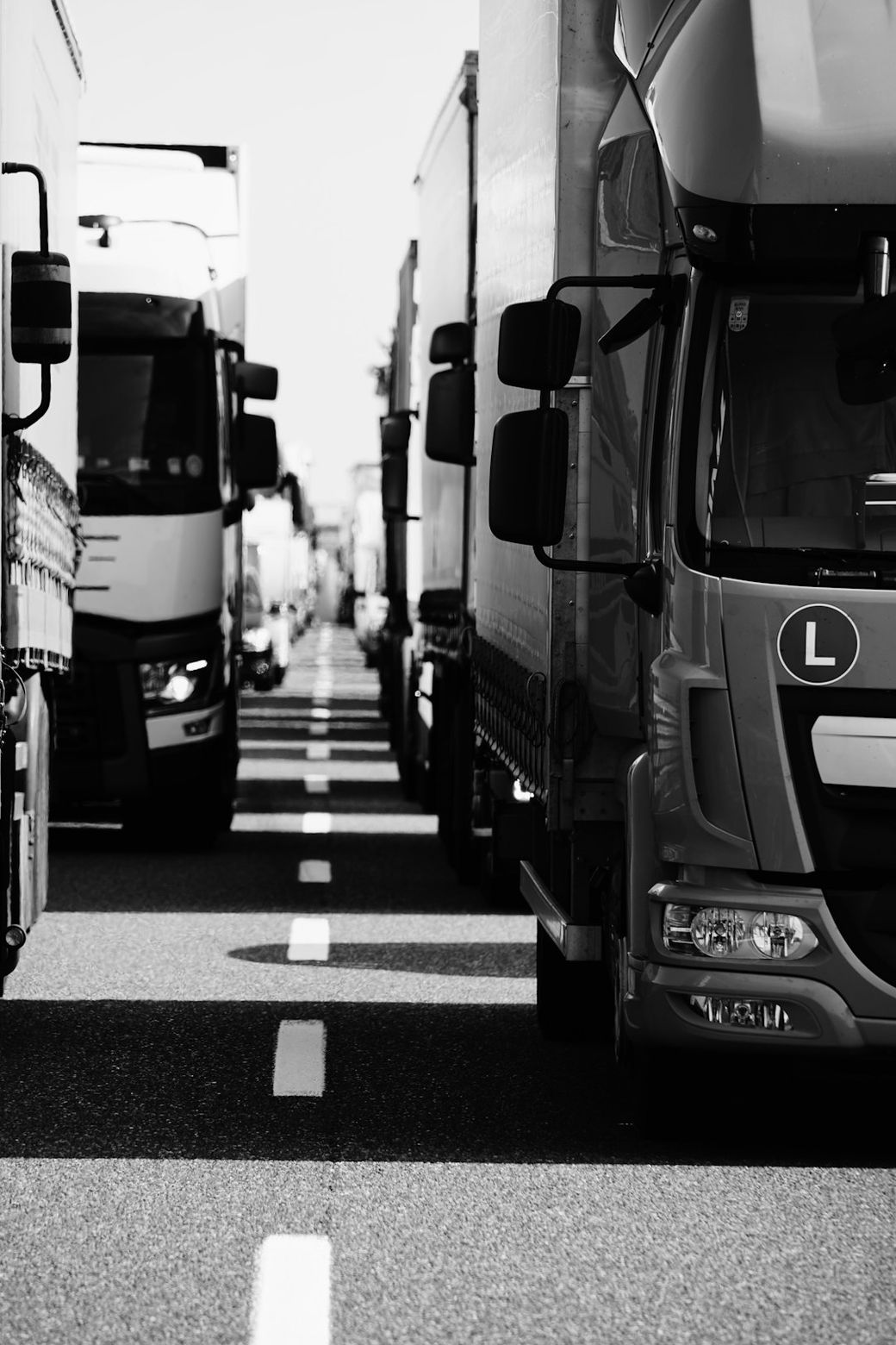 The image features a line of heavy trucks parked closely on a road, creating a narrow pathway between them. The scene is depicted in black and white, emphasizing the shapes and textures of the vehicles. — Credit: Photo by Wolfgang Hasselmann on Unsplash
