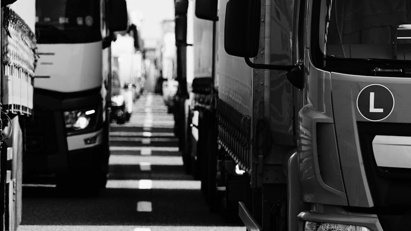 The image features a line of heavy trucks parked closely on a road, creating a narrow pathway between them. The scene is depicted in black and white, emphasizing the shapes and textures of the vehicles.