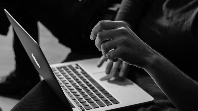 A close-up of a person's hand interacting with a laptop keyboard, with another hand raised in a thoughtful gesture. The image captures a focused moment in a working or studying environment.