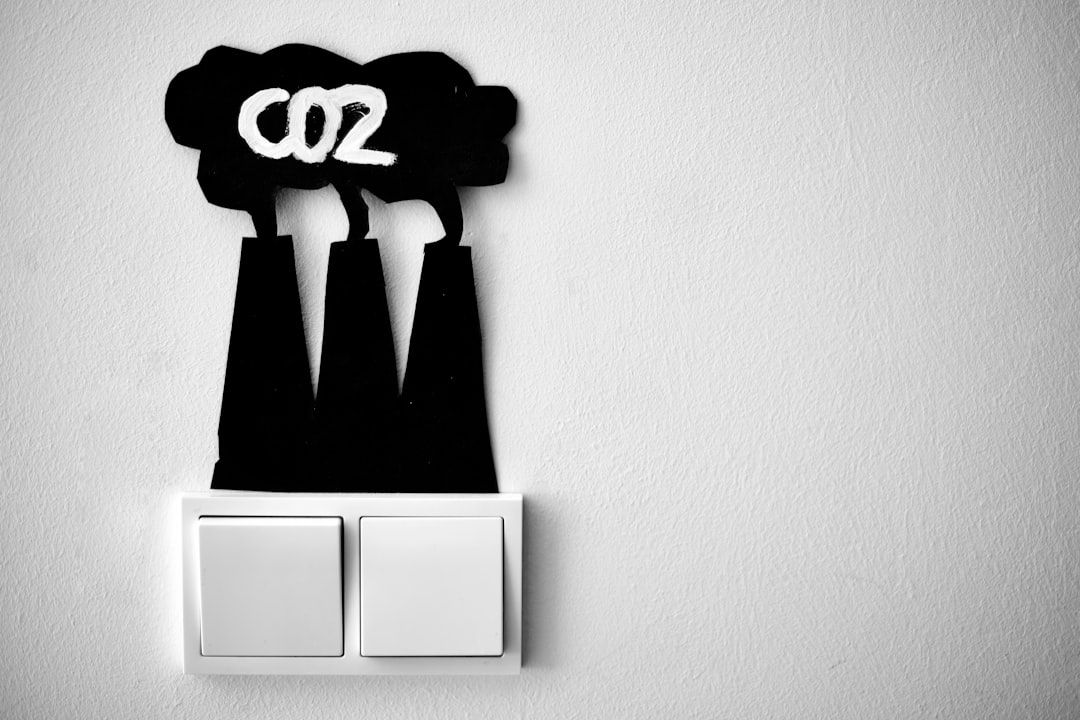 The image depicts a black silhouette of factory smokestacks with the text 'CO2' written on them, placed near two light switches on a white wall. The contrast between the dark silhouette and the light wall emphasizes the theme of carbon emissions.