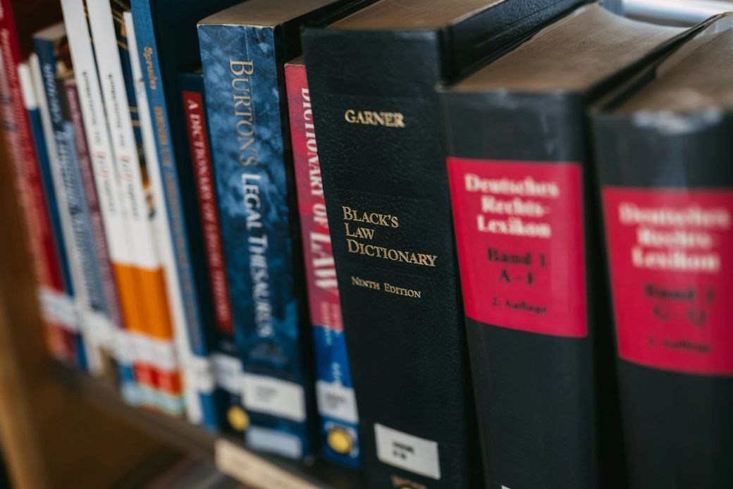 The image shows a close-up of various legal and reference books arranged on a shelf. Prominent among them is 'Black's Law Dictionary' in a black cover, indicating its significance in legal studies. — Credit: Photo by Krists Luhaers / Unsplash