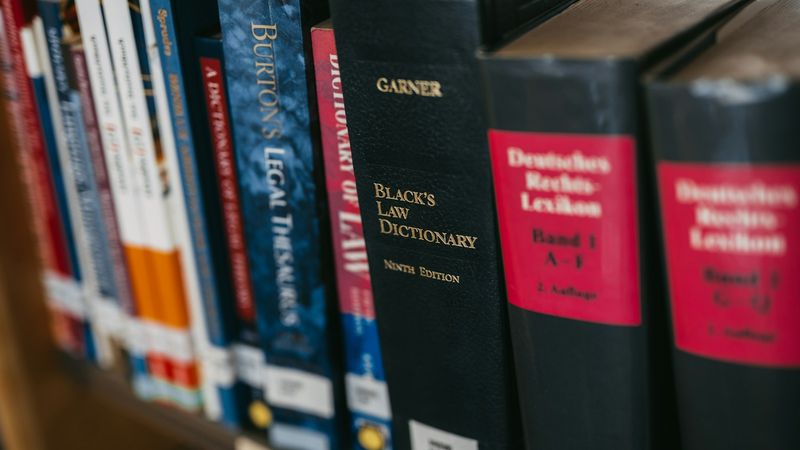 The image shows a close-up of various legal and reference books arranged on a shelf. Prominent among them is 'Black's Law Dictionary' in a black cover, indicating its significance in legal studies.