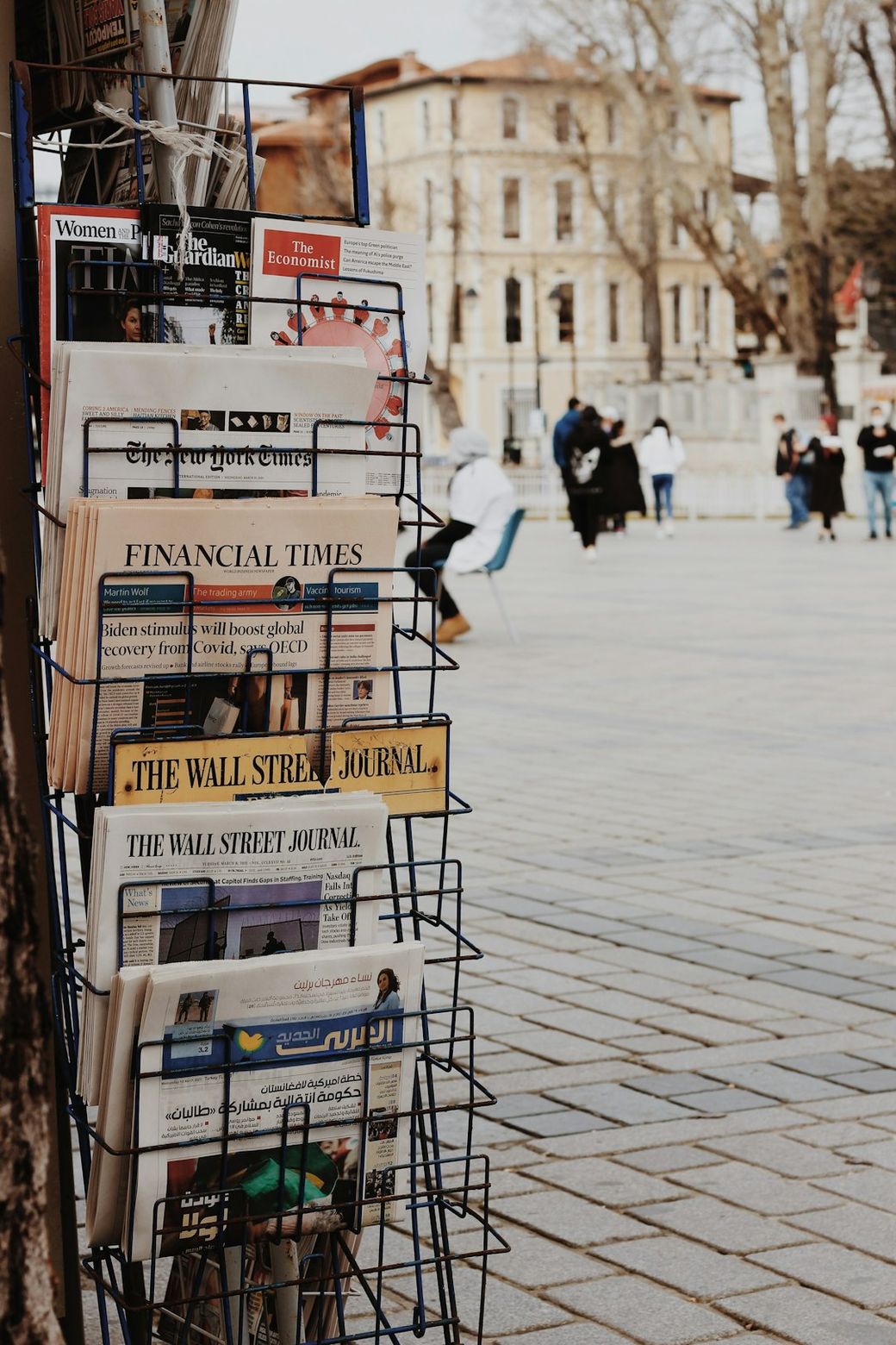 An outdoor newspaper stand displays various newspapers prominently, including 'Financial Times,' 'The Wall Street Journal,' and 'The Economist.' In the background, people are seen walking past buildings in a public square. — Credit: Photo by Hümâ H. Yardım on Unsplash