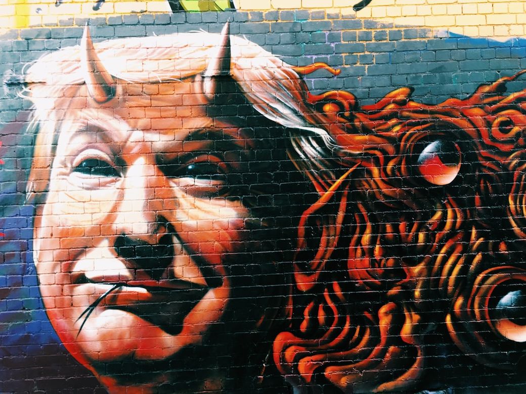 The image depicts a mural of a caricatured figure resembling a former political leader, featuring exaggerated facial features and demonic attributes such as horns and sharp teeth. The artwork showcases vibrant colors against a textured brick wall, conveying a provocative sentiment. — Credit: Photo by Jon Tyson on Unsplash