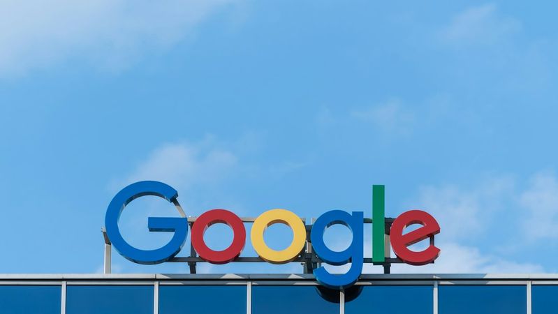 The image features the prominent Google logo displayed on the exterior of a modern building under a clear blue sky. The colorful insignia showcases the company’s branding, emphasizing its identity in the tech industry.