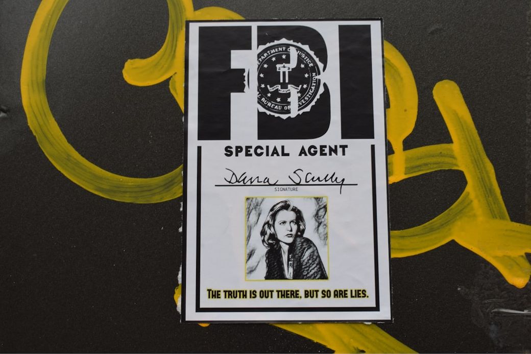 The image features a printed FBI special agent badge with a portrait of a woman named Dana Scully. The badge is affixed to a surface that has yellow graffiti, juxtaposing a professional emblem with urban street art. — Credit: Photo by Marija Zaric on Unsplash