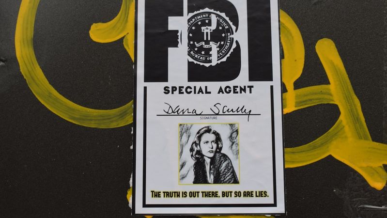 The image features a printed FBI special agent badge with a portrait of a woman named Dana Scully. The badge is affixed to a surface that has yellow graffiti, juxtaposing a professional emblem with urban street art.