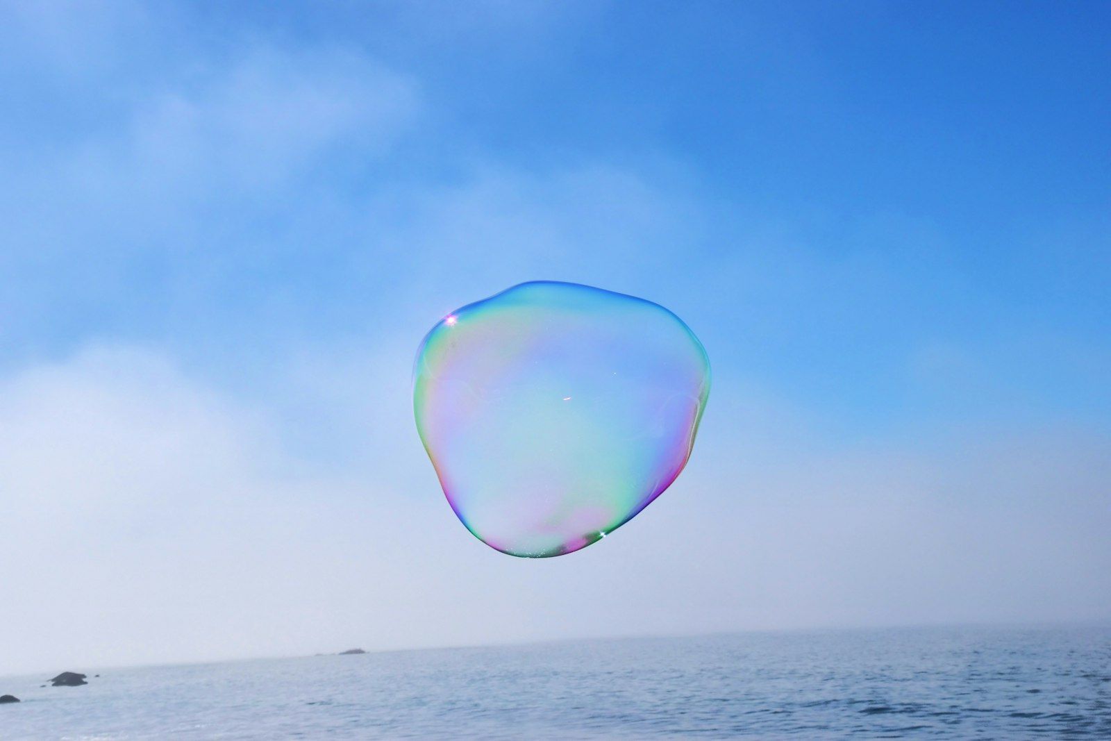 A colorful soap bubble floats in the air above a serene body of water, with a clear blue sky in the background. The bubble reflects a spectrum of colors as it hovers, creating a whimsical and light-hearted scene.