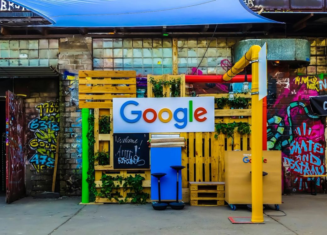 The image features a colorful installation with a prominent Google logo set against a backdrop of graffiti and wooden structures. The scene suggests a creative or tech-related event space designed to attract visitors. — Credit: Photo by Rajeshwar Bachu / Unsplash