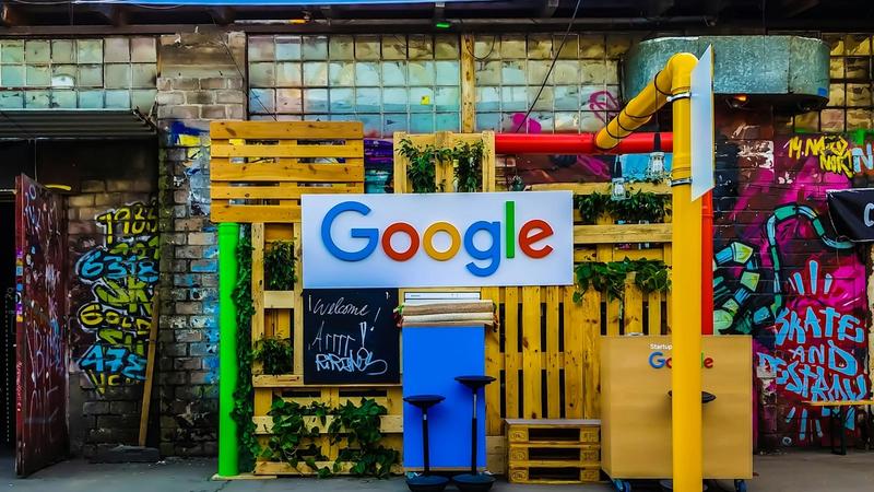 The image features a colorful installation with a prominent Google logo set against a backdrop of graffiti and wooden structures. The scene suggests a creative or tech-related event space designed to attract visitors.