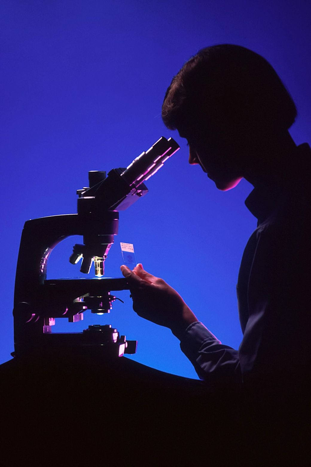 The image features the silhouette of a person looking through a microscope against a vibrant blue and purple background. The focus is on the microscope, highlighting its intricate design and function in scientific exploration. — Credit: Photo by National Cancer Institute / Unsplash