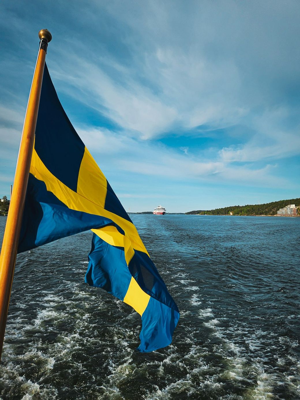 The image captures a Swedish flag waving in the foreground as a cruise ship sails along a waterway, with a scenic landscape in the background. The sky is clear and blue, reflecting a calm atmosphere. — Credit: Photo by Evangelos Vamvounakis on Unsplash