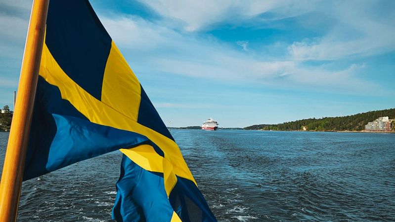 The image captures a Swedish flag waving in the foreground as a cruise ship sails along a waterway, with a scenic landscape in the background. The sky is clear and blue, reflecting a calm atmosphere.