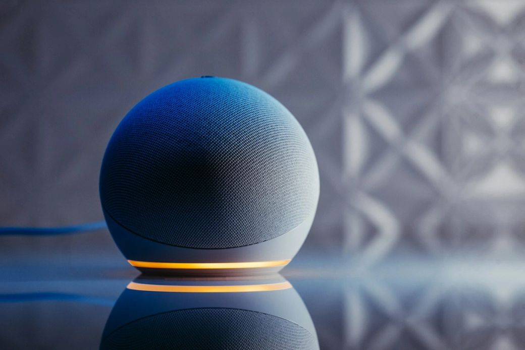 The image features a modern smart speaker with a spherical design, illuminated by a soft orange light. The sleek surface reflects the speaker's design against a textured background. — Credit: Photo by Jonathan Borba / Unsplash