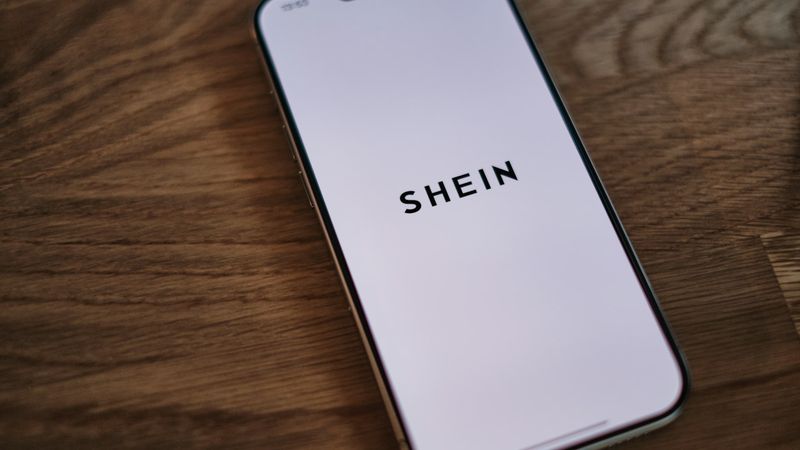 A close-up image of a smartphone displaying the logo of Shein on its screen. The device rests on a wooden surface with a neutral background.