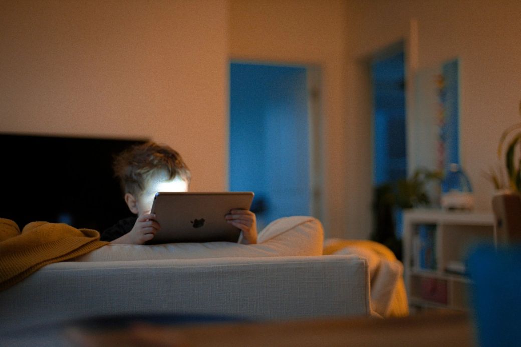 A child is sitting on a couch, illuminated by the screen of a tablet they are holding. The warm lighting creates a cozy atmosphere in the room, contrasting with the cooler tones visible in the background. — Credit: Photo by Nils Huenerfuerst / Unsplash