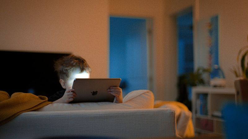 A child is sitting on a couch, illuminated by the screen of a tablet they are holding. The warm lighting creates a cozy atmosphere in the room, contrasting with the cooler tones visible in the background.