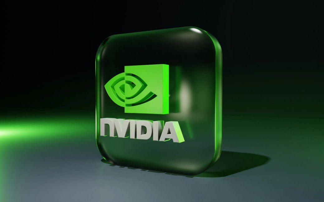 The image features a 3D rendering of the NVIDIA logo on a reflective surface, set against a dark backdrop with green highlights. The logo includes a stylized eye and is predominantly represented in a green and white color scheme. — Credit: Photo by BoliviaInteligente / Unsplash