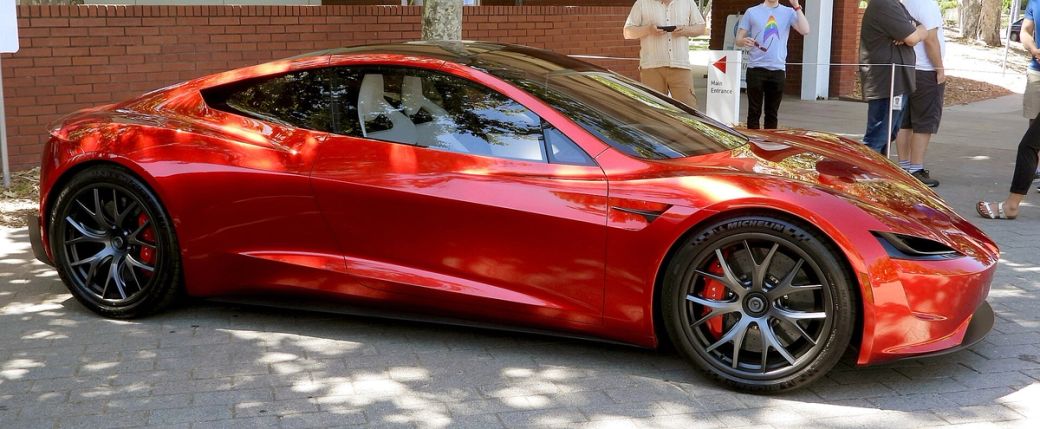 The image showcases a sleek, red Tesla Roadster parked on a brick surface, highlighting its aerodynamic design and modern aesthetic. The bright sunlight enhances the car's glossy finish, while passersby in the background indicate a public event or exhibition setting.