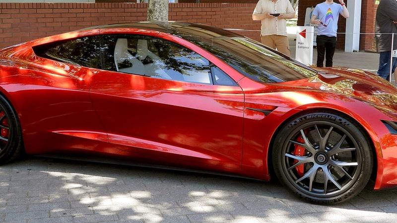 The image showcases a sleek, red Tesla Roadster parked on a brick surface, highlighting its aerodynamic design and modern aesthetic. The bright sunlight enhances the car's glossy finish, while passersby in the background indicate a public event or exhibition setting.
