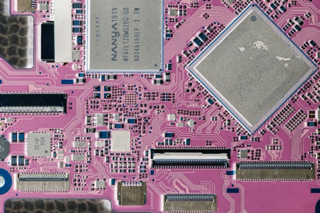 The image displays a close-up view of a pink circuit board, featuring various electronic components such as microchips and capacitors. The intricate patterns and connections are indicative of advanced technology in electronics. — Credit: Photo by Albert Stoynov / Unsplash