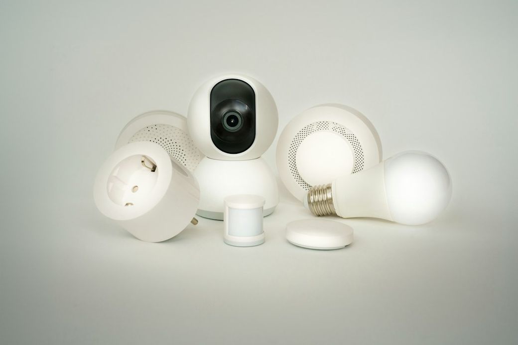 The image features a collection of smart home devices, including a security camera, smart light bulb, motion sensor, and smart plug, all designed in a sleek white aesthetic. These devices are arranged artistically on a neutral background, highlighting their modern technology. — Credit: Photo by Jakub Żerdzicki on Unsplash